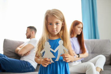 Ein Mädchen steht zwischen getrennten Eltern und hält Männlein und Weiblein aus Papier in seinen Händen