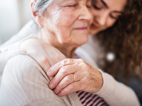 Eine Seniorin hält die Hand einer sie umarmenden jungen Frau