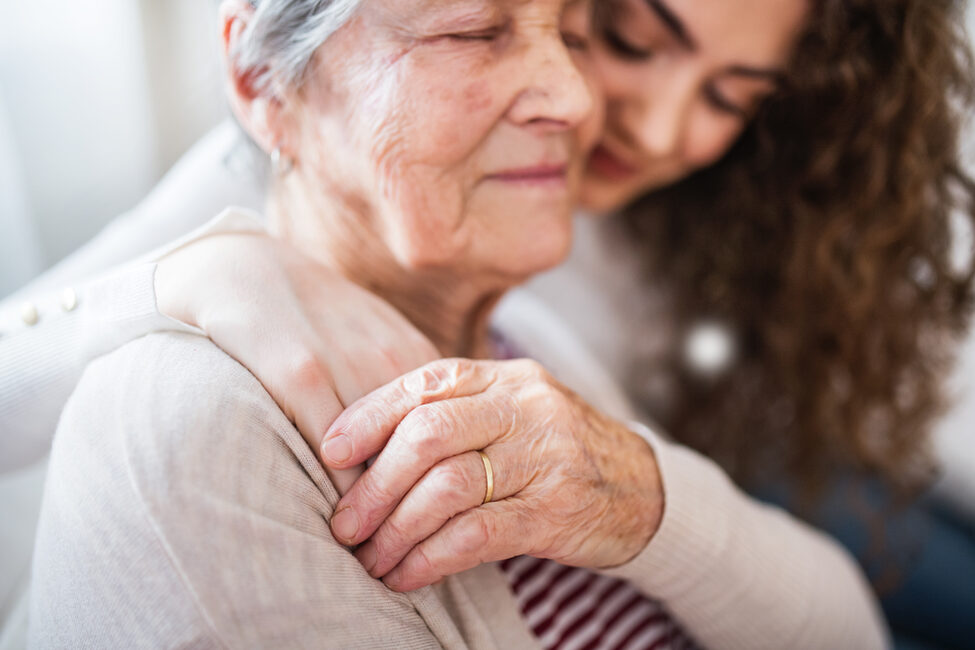 Eine Seniorin hält die Hand einer sie umarmenden jungen Frau