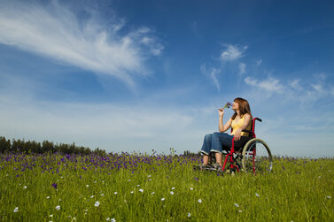 Eine Frau sitzt auf einem Rollstuhl. Sie befindet sich auf einer Wiese und riecht an Blumen.