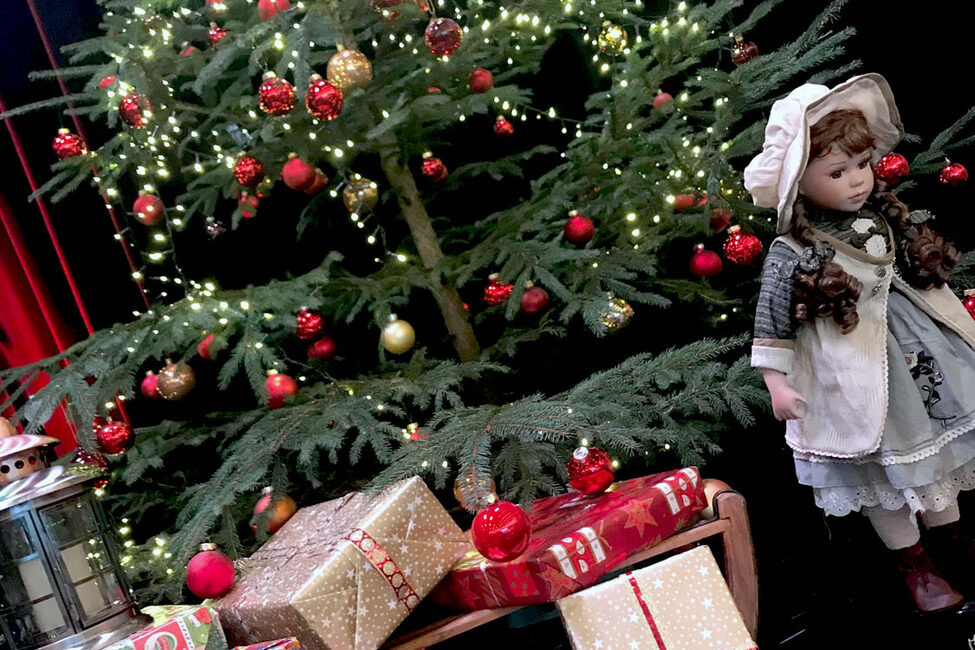 Ein geschmückter Weihnachtsbaum, davor Geschenke und eine Puppe