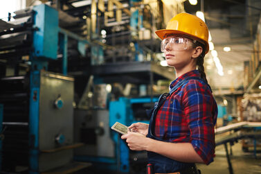 Eine junge Frau hält eine tablet und steuert eine große Maschine in einer Werkstatt