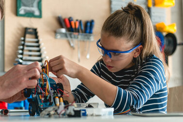 Ein Mädchen baut ein einem kleinen elektrischen Auto, im Hintergrund ist eine Werkstatt zu sehen