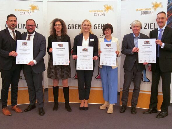 Sieben Personen aus Politik, Ministerium, Regierung und Verwaltung stehen eng beieinander und lachen fröhlich in die Kamera. Sie halten stolz Urkunden in den Händen – ein Moment der gemeinsamen Freude und des Erfolgs.