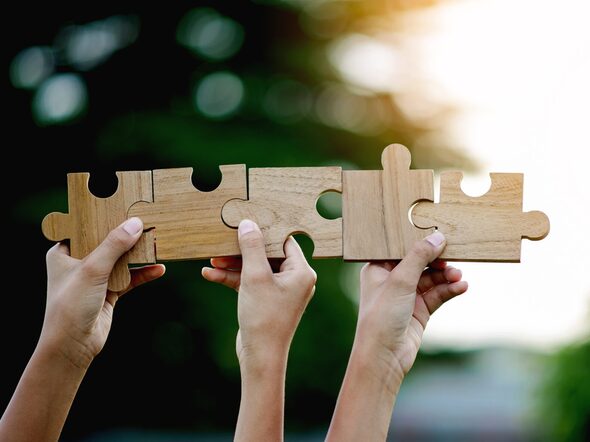 Fünf Holzpuzzleteile in einer Reihe, die von drei Händen in die Luft gehalten werden.