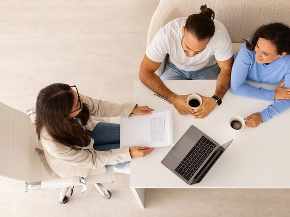Luftbild von oben auf drei Personen an einem Tisch mit Laptop, Block und Kaffee.