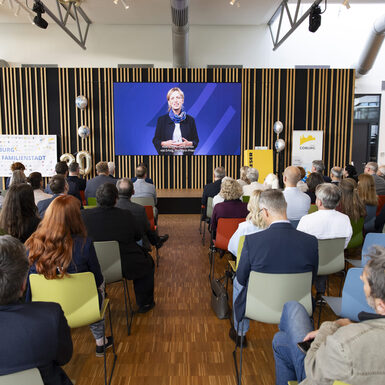 Publikum sitzt auf Stühlen vor der Bühne und schaut auf eine Leinwand an der Bühne.