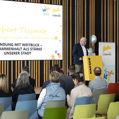 Publikum sitzt auf Stühlen vor der Bühne. Ein Mann steht am Rednerpult auf der Bühne.