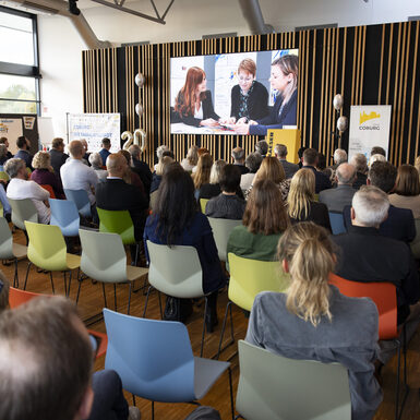 Publikum sitzt auf Stühlen vor der Bühne und schaut auf eine Leinwand an der Bühne.