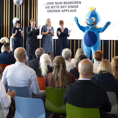 Publikum sitzend auf Stühlen vor der Bühne. Auf der Bühne stehen mehrere Personen und ein Maskottchen.