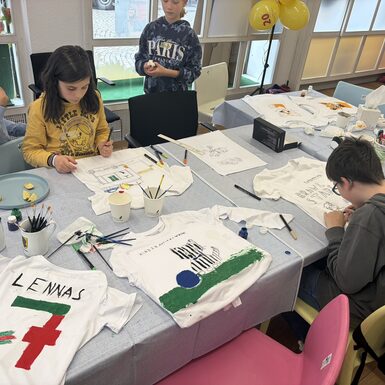 Kinder stehen und sitzen um einen Tisch und bemalen T-Shirts.