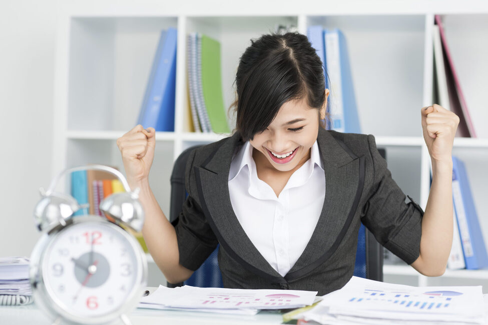 Junge Frau am Schreibtisch mit Unterlagen und Wecker. Sie streckt die Fäuste nach oben.