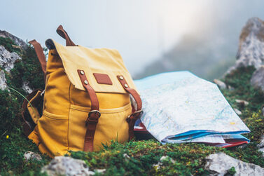 Ein gelber Wanderrucksack und eine Landkarte liegen auf einem Felsen. Im Hintergrund zeichnet sich eine Berglandschaft ab.