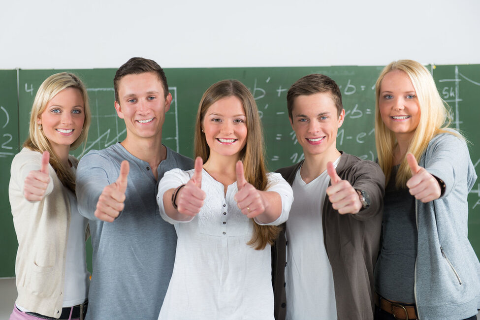 Fünf lächelnde Jugendliche stehen in einem Klassenraum vor der Tafel und zeigen