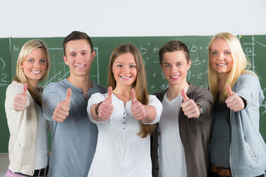 Fünf lächelnde Jugendliche stehen in einem Klassenraum vor der Tafel und zeigen